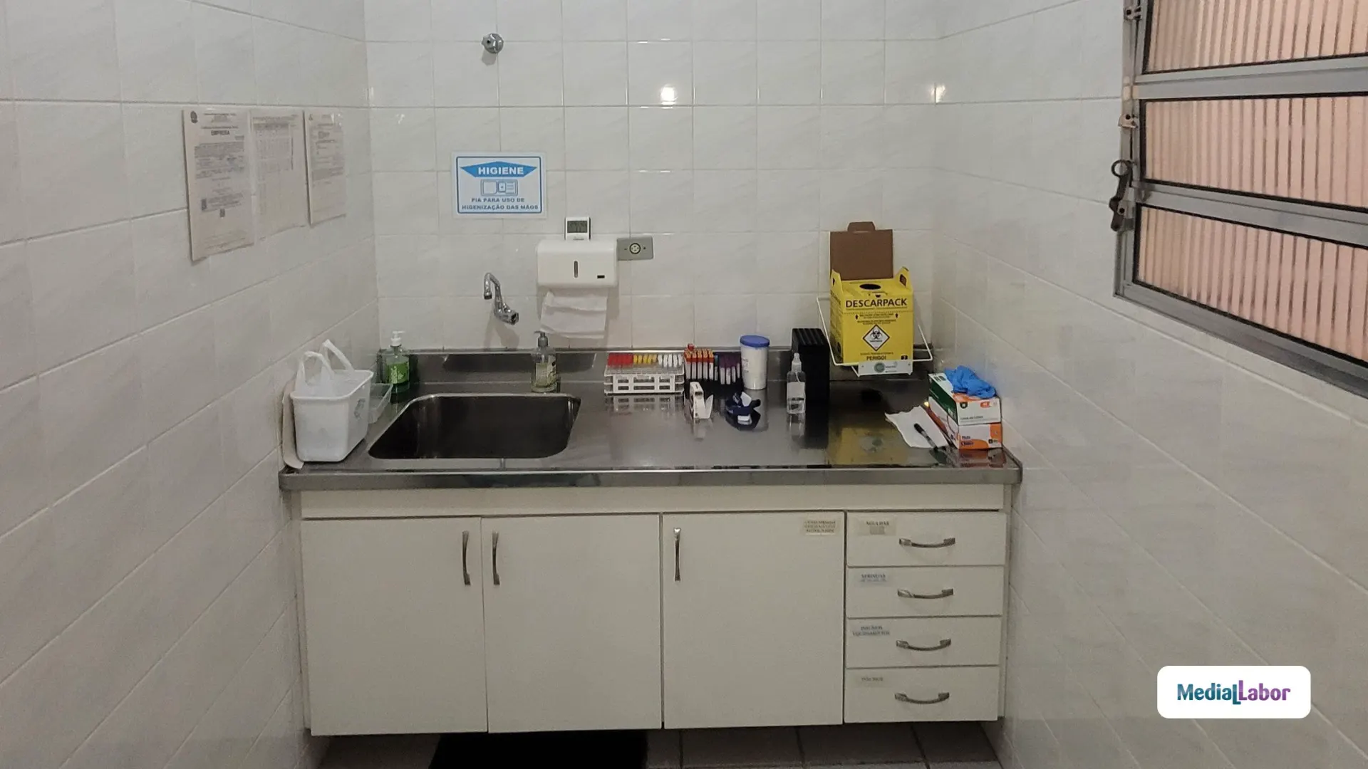 Pia de aço inox com torneira e bancada em laboratório, com dispenser de higiene, organizadores e materiais sobre a bancada em ambiente com azulejos claros.