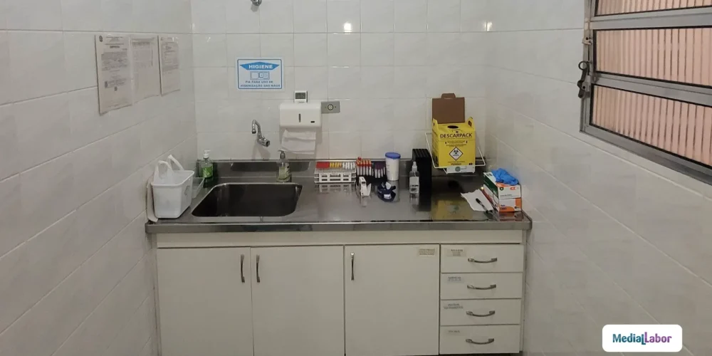 Pia de aço inox com torneira e bancada em laboratório, com dispenser de higiene, organizadores e materiais sobre a bancada em ambiente com azulejos claros.